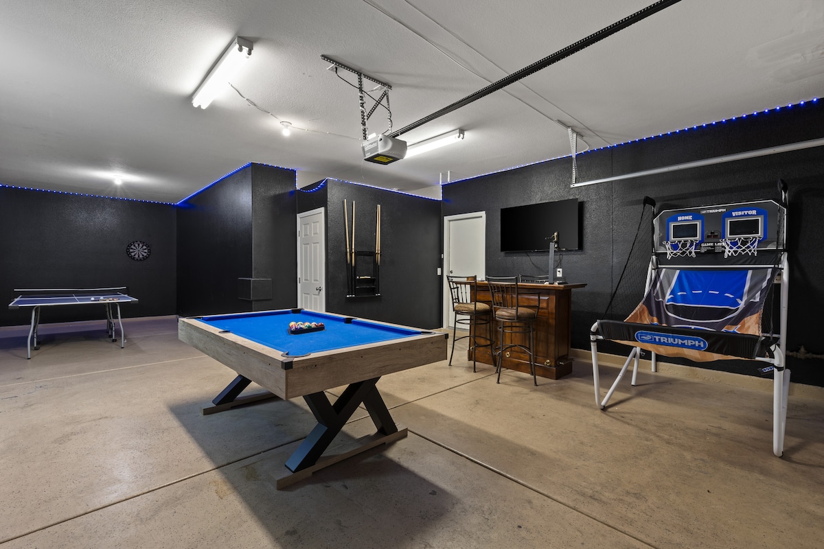 An inviting game room features a blue pool table, alongside a ping-pong table, a mini basketball arcade game, and a dart board. A bar area with seating is visible, and a 55" TV is mounted on the wall, creating a lively entertainment space.