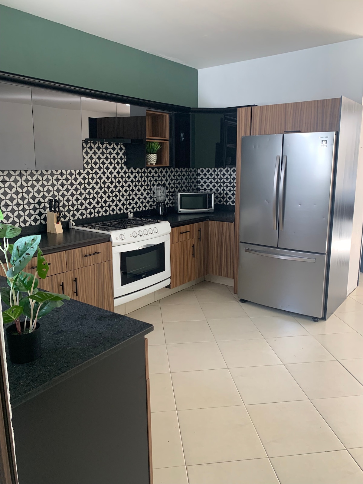 A modern kitchen is presented with sleek cabinetry in earthy tones and a striking geometric tile backsplash. The space features essential appliances, including a stainless steel refrigerator and an oven, with ample counter space complemented by a small potted plant on the counter.