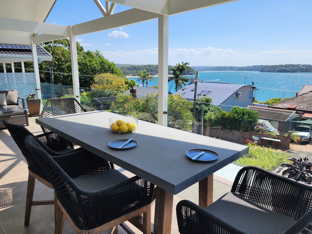 An outdoor dining area features a large concrete table with seating for six. Two plates are set on the table, accompanied by a bowl of lemons. A stunning view of the water and surrounding greenery is visible in the background.