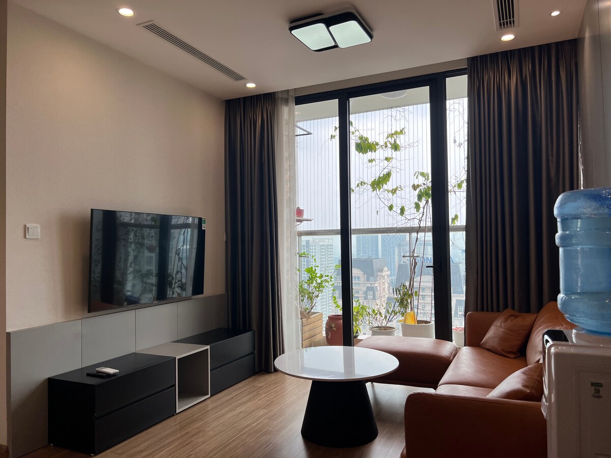 A modern living area is presented with a comfortable brown sofa and a round coffee table positioned in front of a large flat-screen TV. Floor-to-ceiling windows allow natural light to fill the space, showcasing a view of cityscape and indoor plants on the balcony.