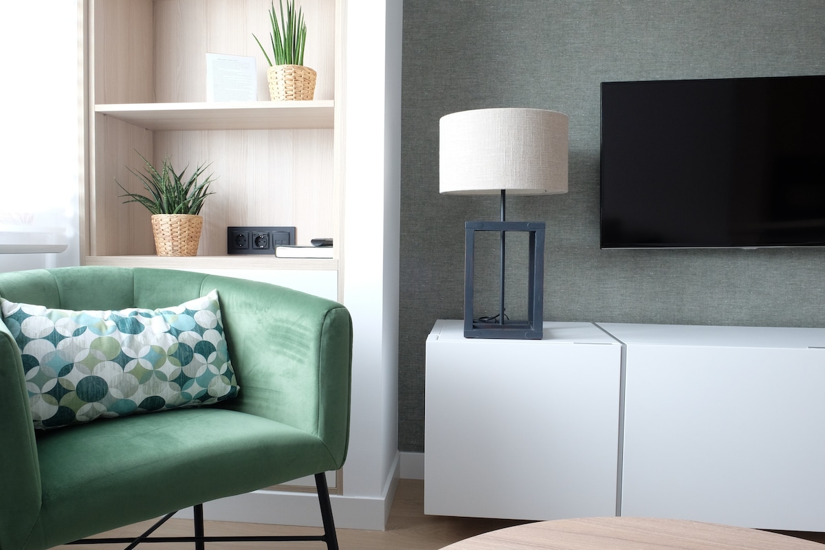 A modern living space is showcased, featuring a green, plush armchair beside a light wooden shelf accented with potted plants. A flat-screen TV is mounted on the wall above a sleek white unit, complemented by a stylish lamp with a fabric shade.