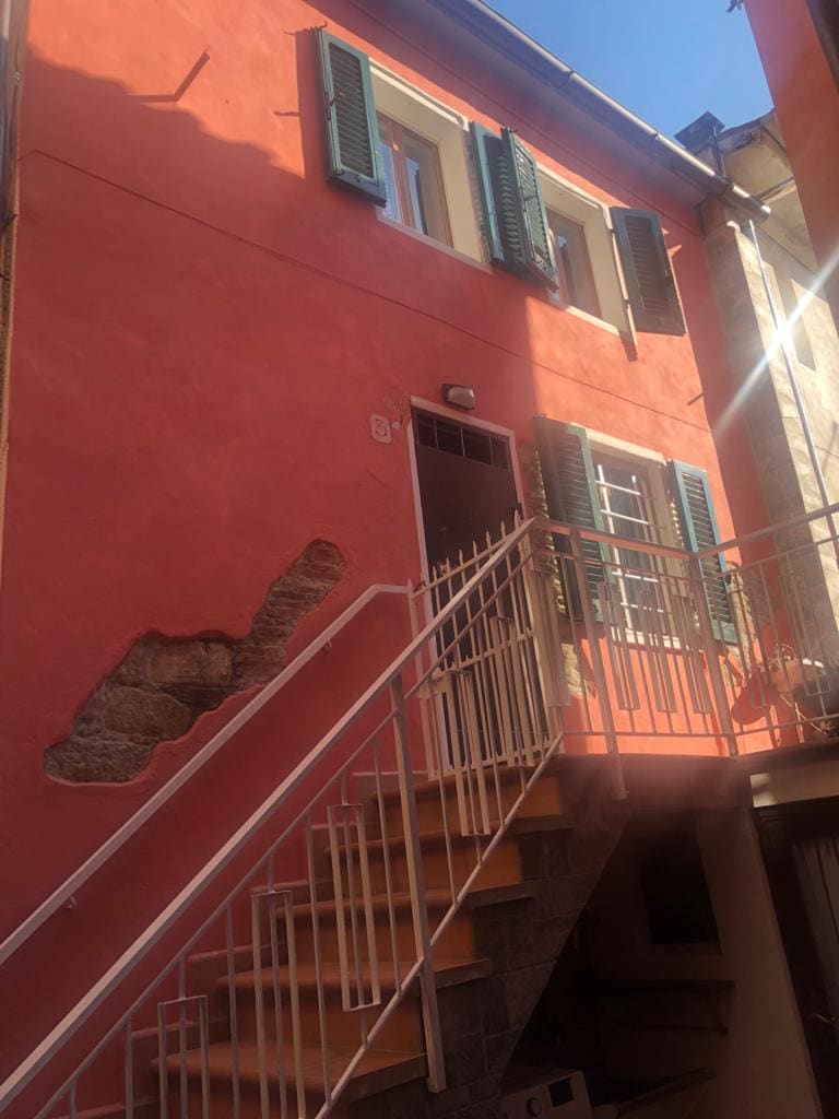 The exterior of a charming two-story building features textured pink walls and green shutters. A metal staircase leads to the entrance, with light illuminating the façade and highlighting the details of the architecture.