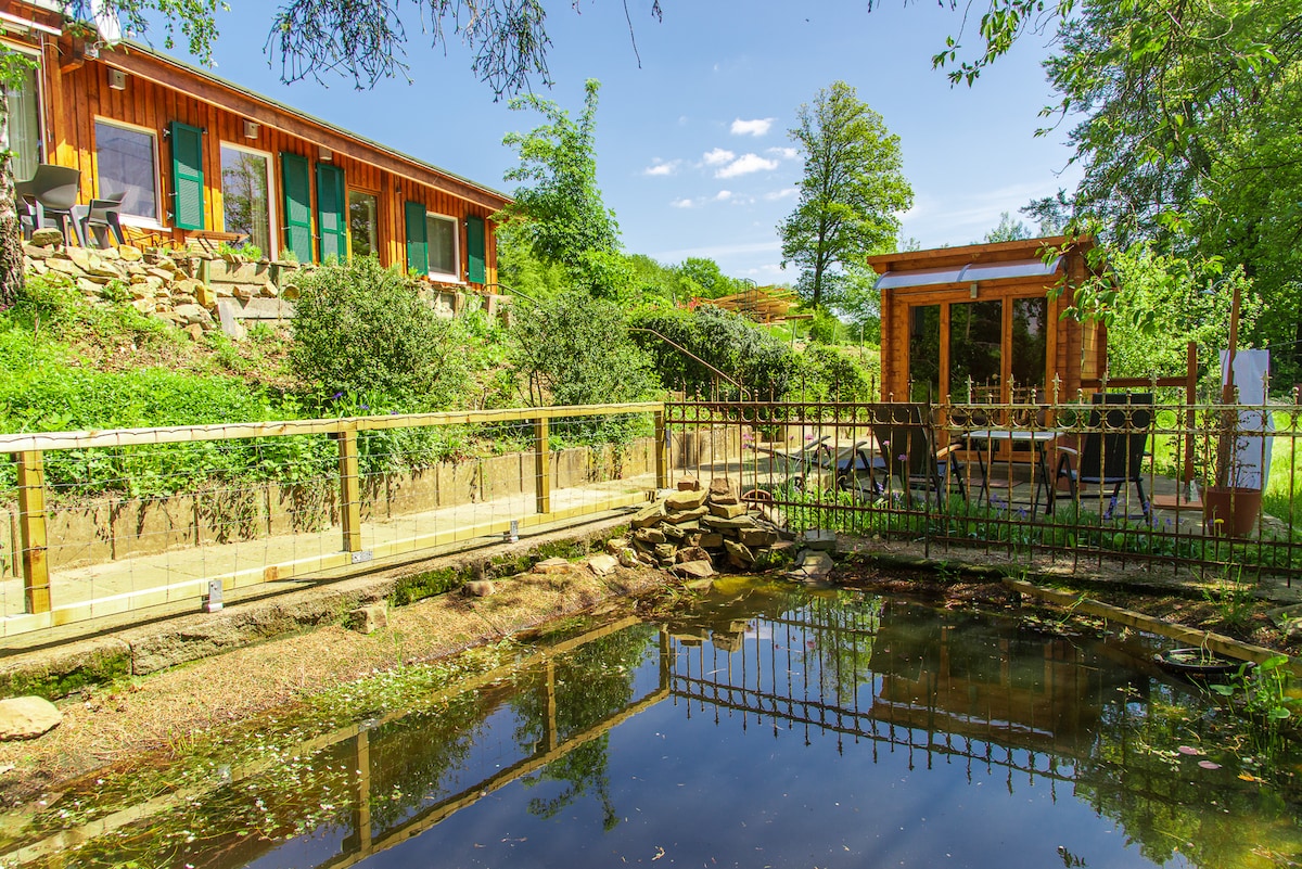 A serene garden scene showcases a tranquil pond reflecting the surrounding greenery. Nearby, a wooden lodge is visible with large windows and a wraparound deck, while a small sauna structure is nestled among the trees in the background.