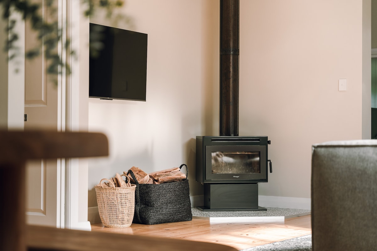 A living area features a combustion fireplace set against a neutral wall. Relaxed seating is positioned nearby, while a basket of firewood adds a cozy touch. A flat-screen TV is mounted on the wall, contributing to the inviting ambiance.