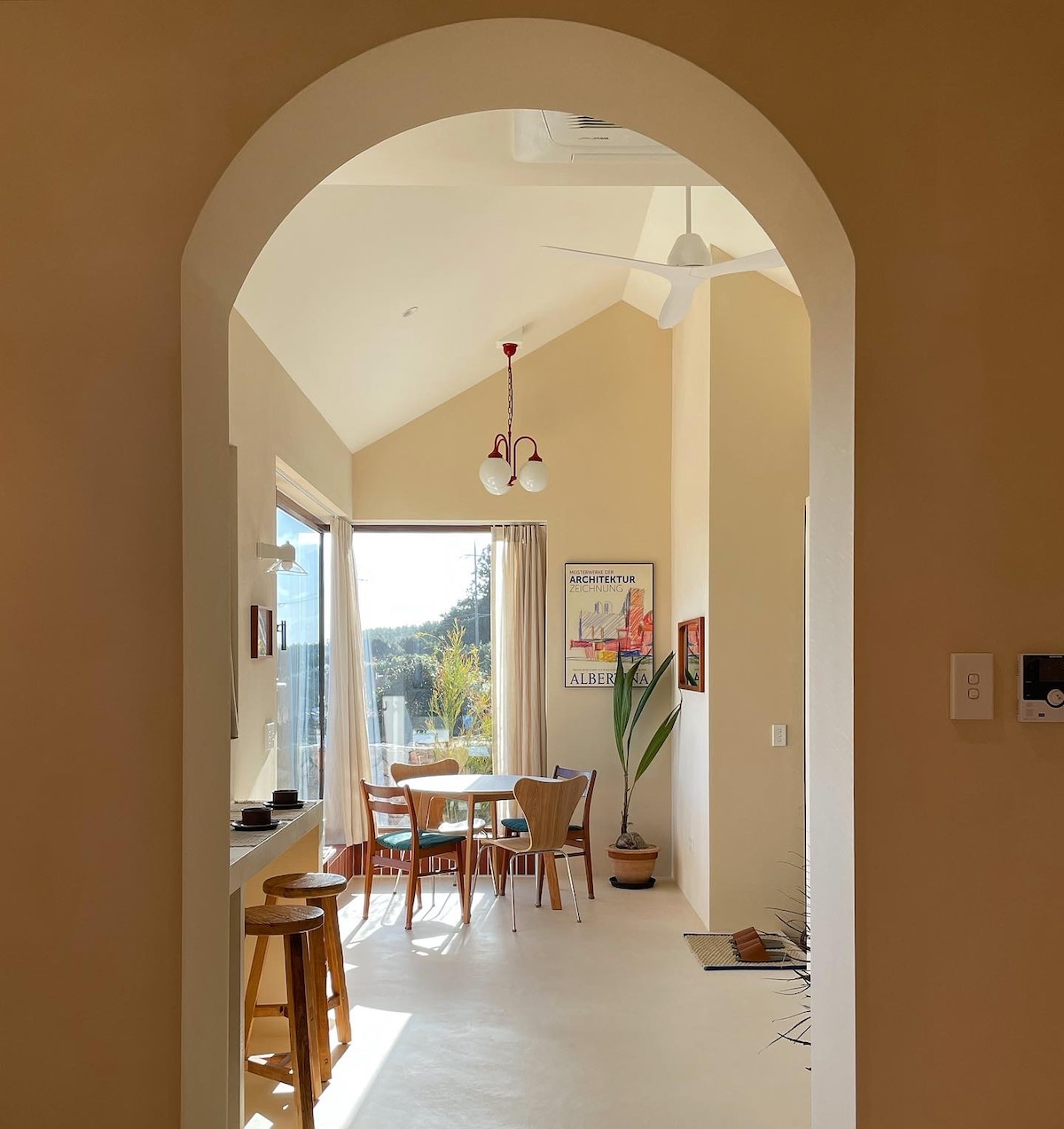 An arched entryway provides a view into a dining area, featuring a table surrounded by chairs. Natural light fills the space through large windows, highlighting the warm tones of the walls and a potted plant adding a touch of greenery.