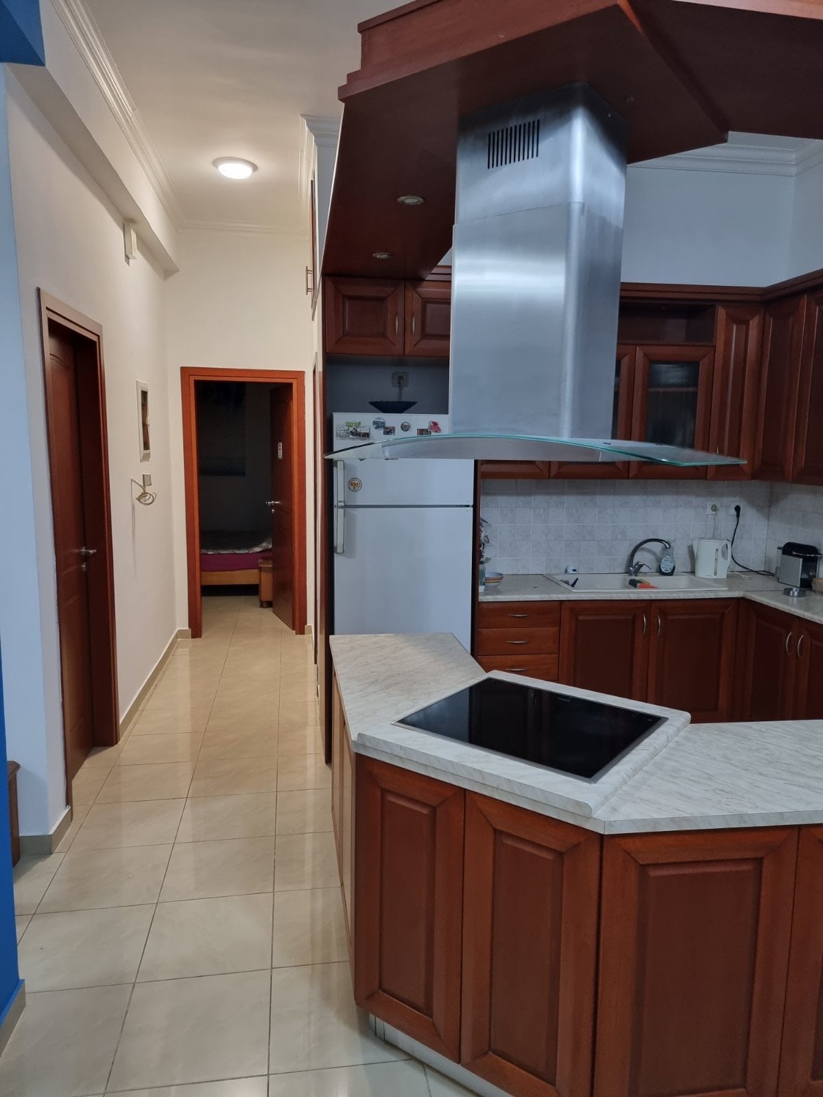 A functional kitchen features rich wooden cabinetry and a sleek countertop. An island with an integrated cooktop is positioned centrally. The room connects to a hallway leading to other areas of the home, with soft lighting enhancing the efficient layout.