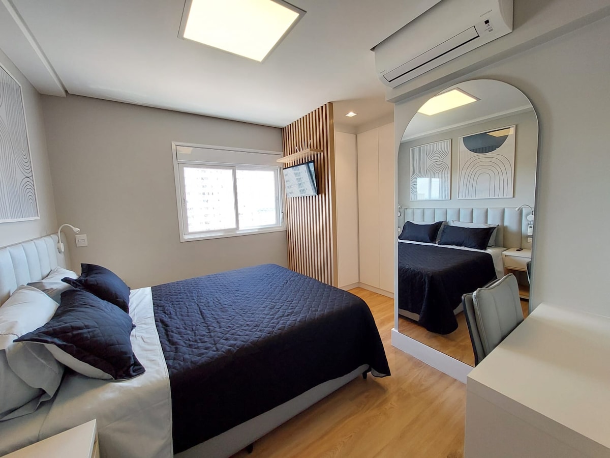 A contemporary bedroom is featured, equipped with a spacious bed dressed in dark blue and light blue linens. A wall-mounted TV is positioned in front of the bed. A large mirror enhances the space, reflecting natural light from the window on the side.