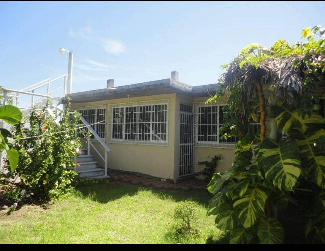 A single-story building is surrounded by vibrant greenery, showcasing a light-colored exterior with large windows secured by metal grilles. A set of stairs leads to a covered entryway, while a well-kept lawn adds to the natural setting.