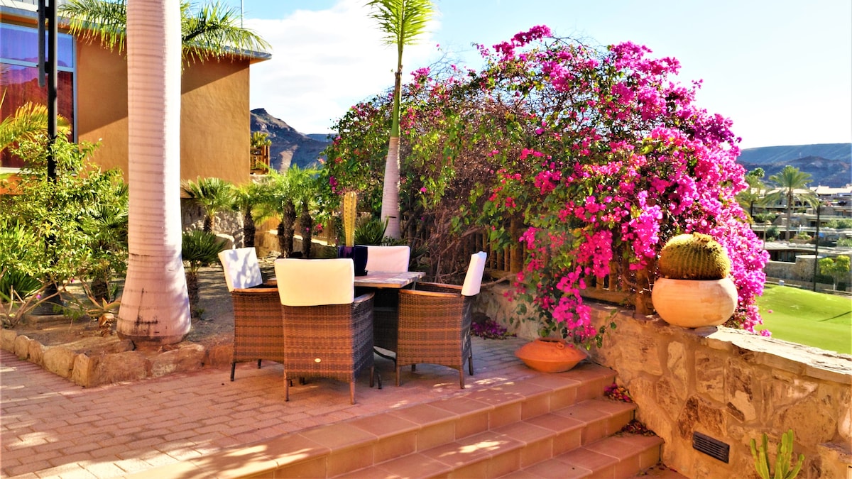 An outdoor seating area is set with wicker chairs and a table, surrounded by vibrant pink bougainvillea. The space is paved with warm-toned tiles and features lush greenery in the background, contributing to a serene and inviting ambiance.