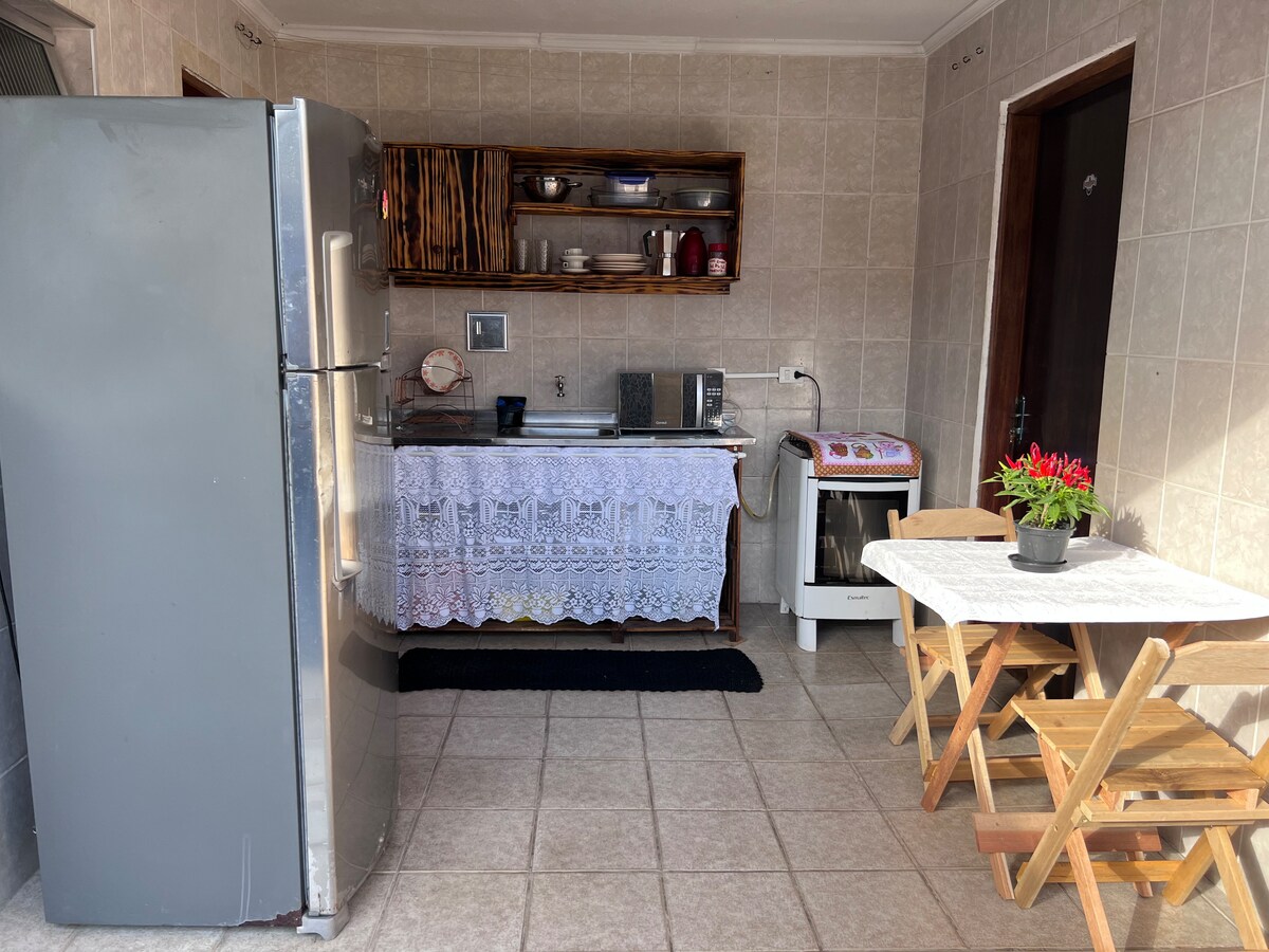 A functional kitchen space is equipped with a large refrigerator, a sink, and a countertop with storage. A small table with folding chairs is positioned next to a potted plant. Light-colored tiles cover the floor, enhancing the room's clean and welcoming appeal.