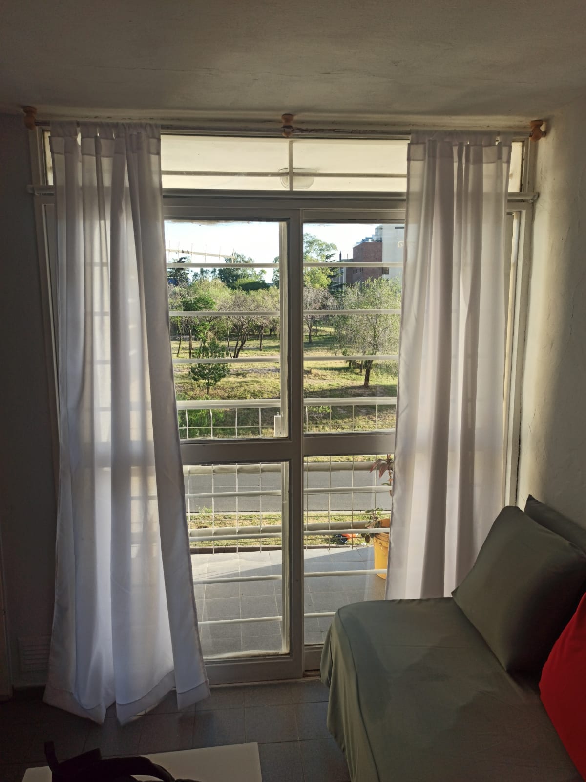 A large window with white curtains allows natural light to flow into the room. The view outside features green trees and open space, creating a sense of tranquility. A comfortable seating area is positioned nearby, enhancing the overall inviting atmosphere.