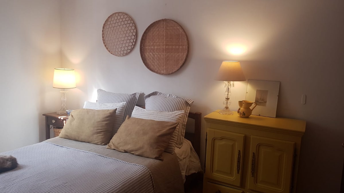 A cozy bedroom features a spacious bed with striped bedding and plush pillows. Two woven wall hangings add texture, while bedside lamps provide warm lighting. A yellow dresser complements the room, enhancing the overall serene atmosphere.