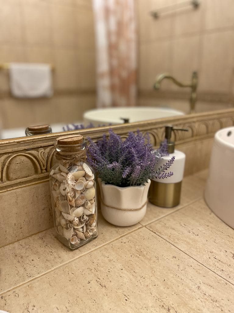 A bathroom countertop is depicted, featuring a glass jar filled with seashells and a small white pot containing lavender. A soap dispenser and a toothbrush holder are present, while a warm-toned mirror reflects the elegant tile design and faucet in the background.