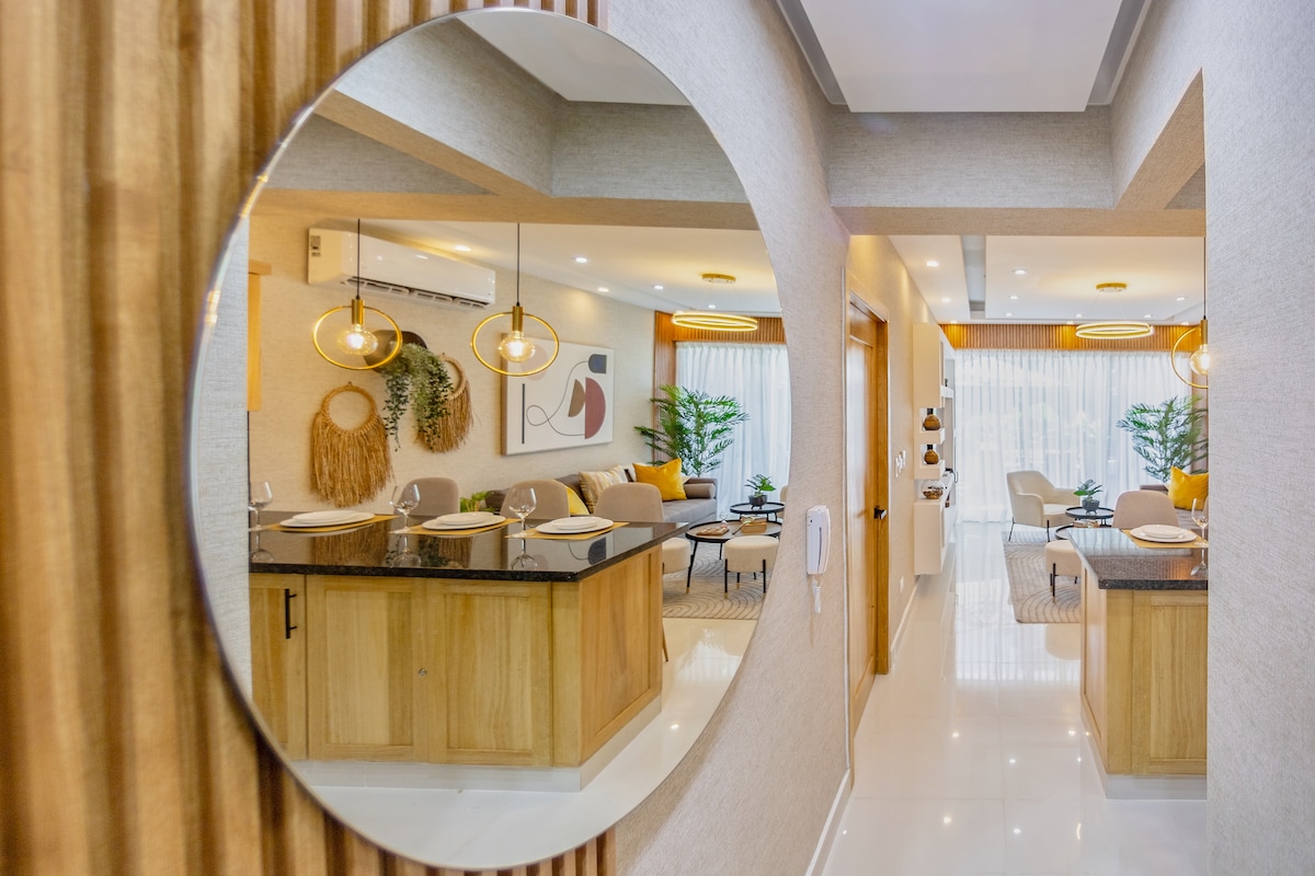 A modern hallway is reflected in a round mirror, showcasing an open living space with stylish furniture and decorative plants. Warm lighting accentuates the welcoming ambiance, while an abstract art piece adorns the wall, enhancing the contemporary design.