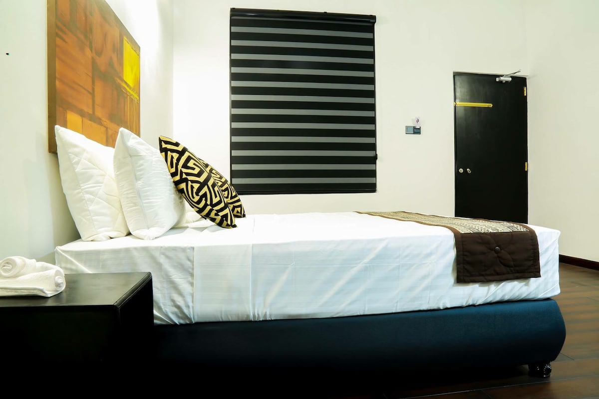 A spacious bed is neatly made with white linens and accented by black and gold patterned pillows. A dark bedside table sits beside the bed. A striped window treatment partially filters natural light, and a black door is visible in the background.