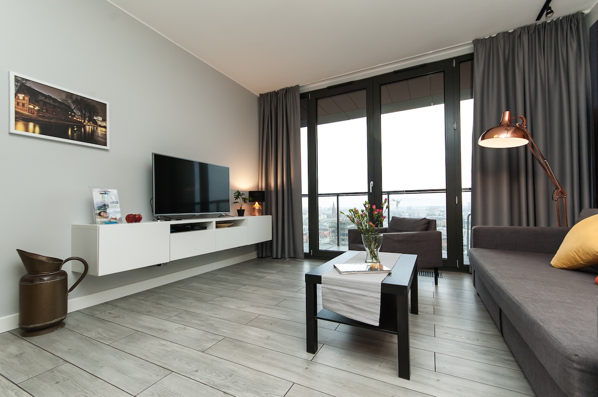 A modern living area is presented with a sleek white entertainment unit and a large flat-screen TV. A comfortable gray sofa and a wooden coffee table are centrally located. Floor-to-ceiling windows are dressed with dark curtains, offering views of the city skyline.