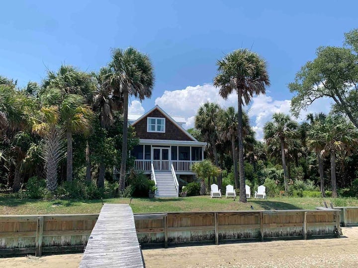A charming elevated cottage is set among palm trees, with a welcoming front porch and steps leading up to it. A wooden walkway extends from the sandy area, leading towards the cottage. The surrounding landscape features lush greenery and is bathed in natural sunlight.
