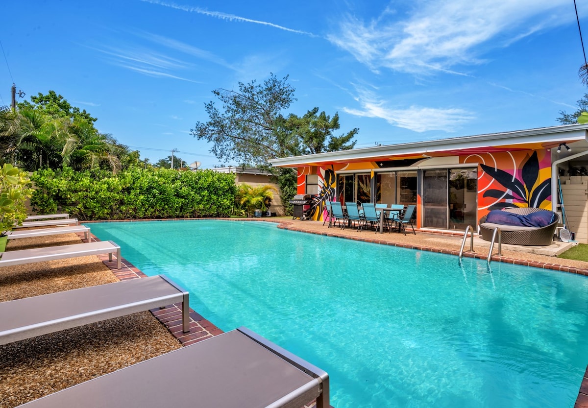 A large outdoor private pool is surrounded by multiple lounge chairs, inviting relaxation. Vibrant wall art decorates the nearby house, which features expansive glass doors leading to the pool area. Lush greenery frames the space, providing a sense of privacy and tranquility.