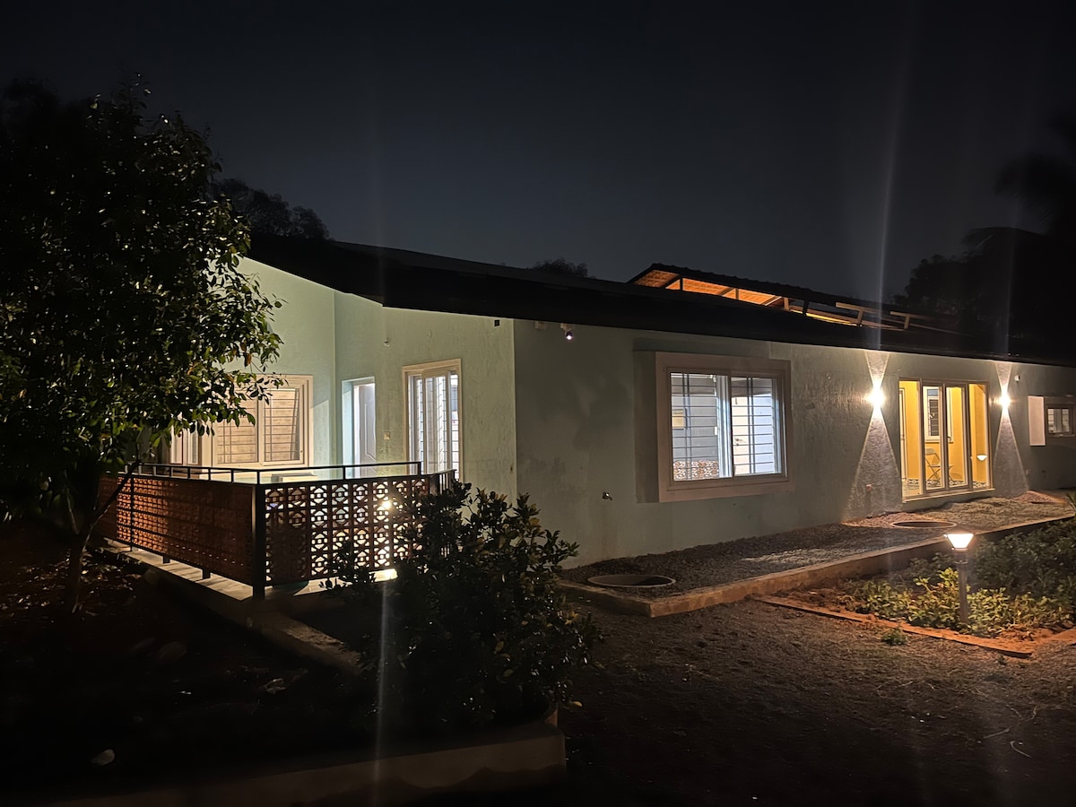 A spacious farmhouse is illuminated at night, showcasing a blend of warm lighting and natural scenery. Large windows reflect the light, creating inviting reflections. The surrounding garden features well-maintained plants and pathways, enhancing the tranquil outdoor setting.