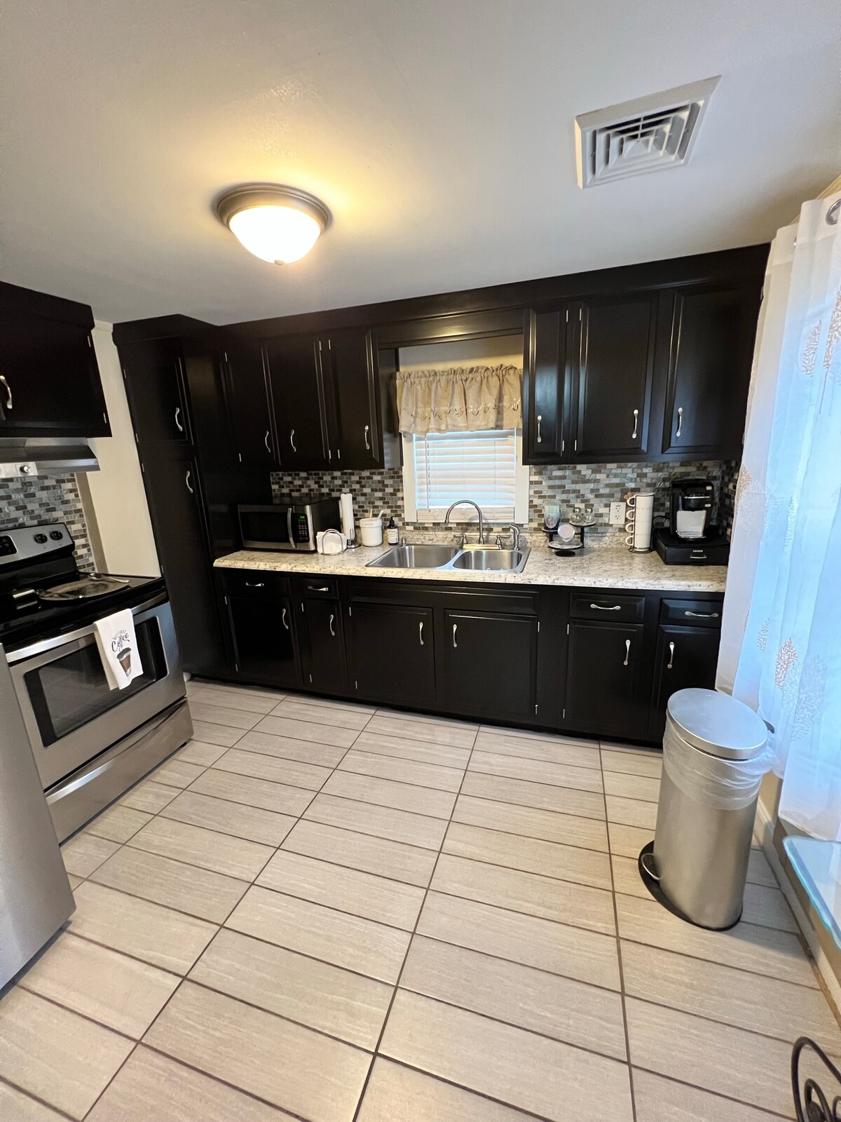 A modern kitchen features dark cabinetry complemented by light-colored countertops. Stainless steel appliances, including a stove and refrigerator, are arranged efficiently. A stainless steel sink is positioned beneath a window with sheer curtains, allowing natural light to brighten the space. Tile flooring is present.