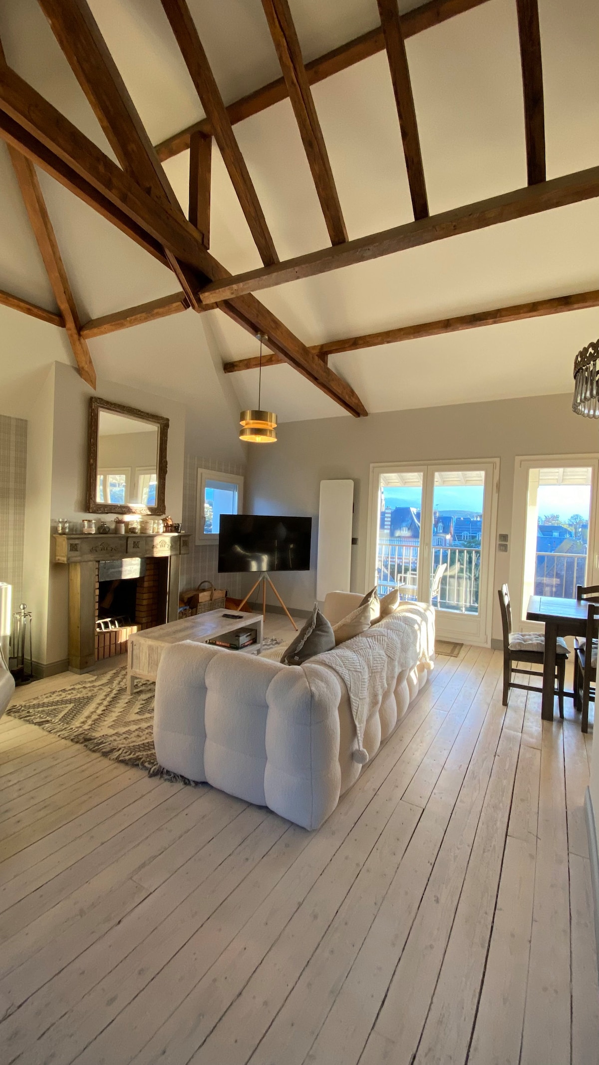 A spacious living area features a high cathedral ceiling with exposed wooden beams. A light-colored sofa faces a modern television, while a cozy fireplace adds character. Large glass doors open to a sunny terrace, enhancing the room's bright ambiance.