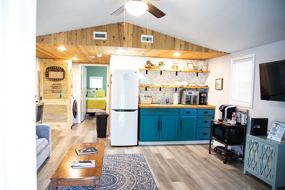 An open living area showcases a kitchenette with teal cabinetry and counter appliances. A cozy seating area is complemented by a coffee station. The space is finished with a wooden ceiling and light-colored walls, creating a bright and airy atmosphere.