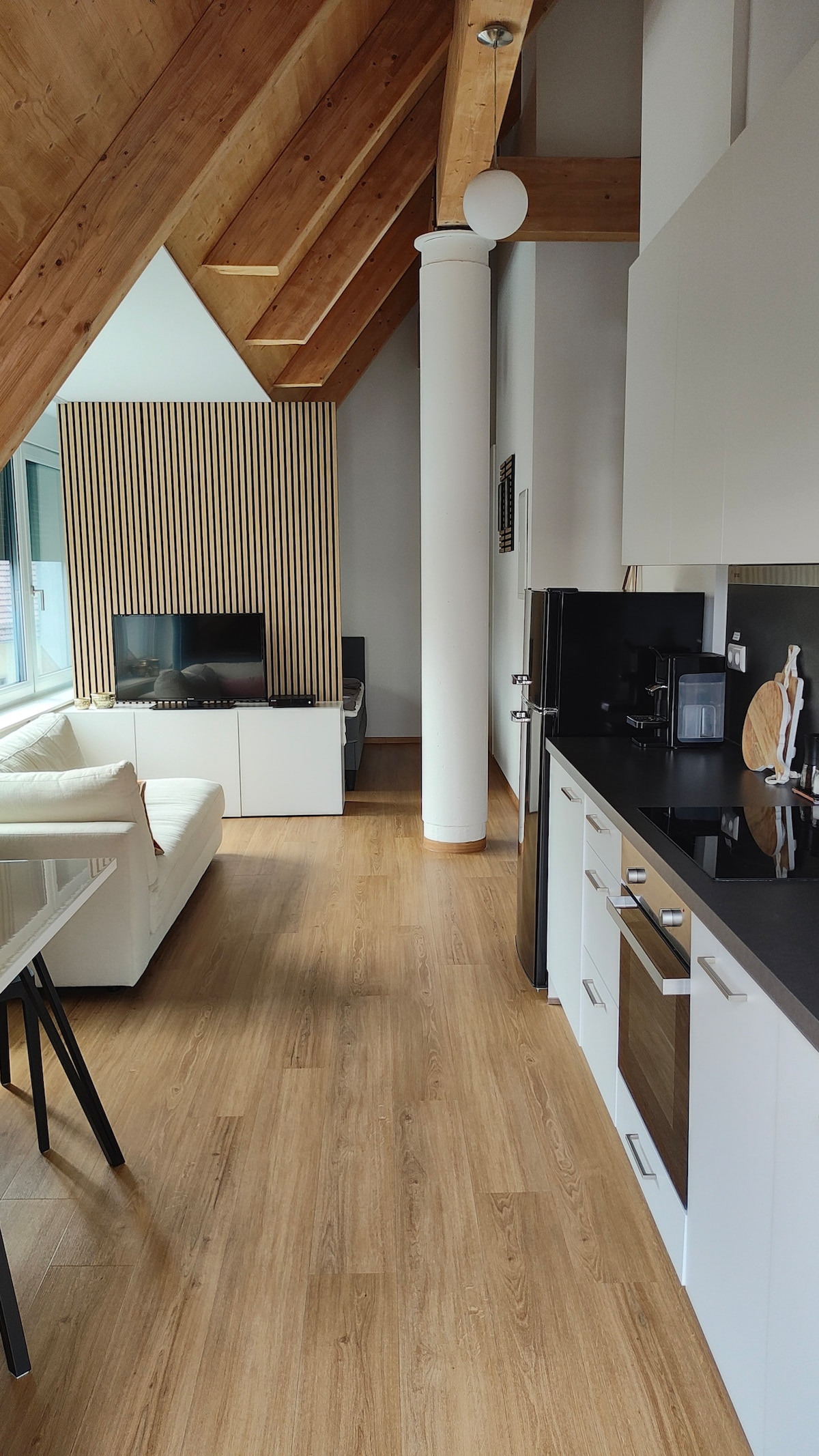 A spacious living area features high wooden ceilings with exposed beams. A sofa faces a TV mounted on a light wood feature wall. To the side, a modern kitchen with a black countertop is equipped with appliances, including an oven and a coffee machine.