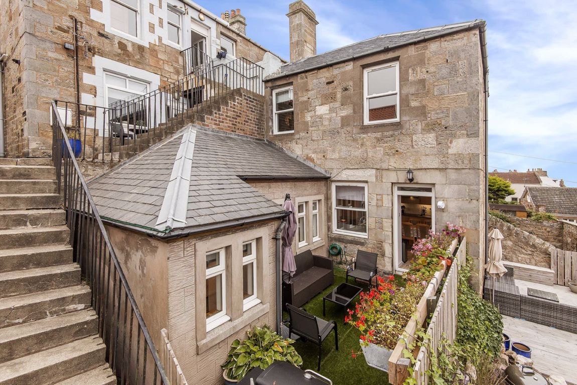 An exterior view of the cottage shows a charming stone structure complemented by a sloped roof and several windows. A private terrace area is visible, featuring outdoor seating and greenery. A staircase leads to an upper level, providing access to additional outdoor spaces.