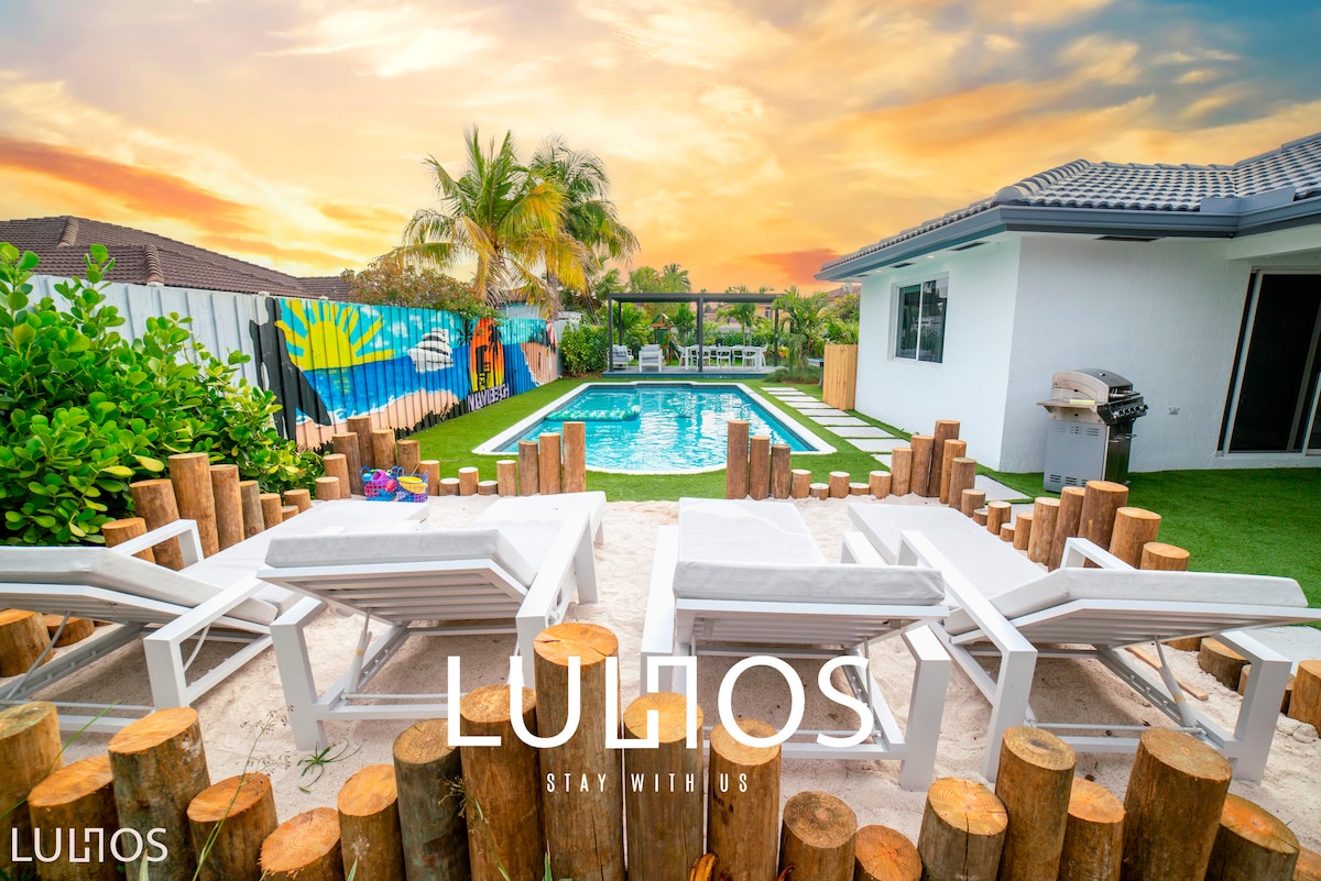 Lounge chairs are arranged on sandy ground, providing a comfortable spot near the heated pool. Lush greenery surrounds the pool area, with colorful beach towels displayed on a fence. The sunset sky fills the background with warm hues, enhancing the outdoor space.