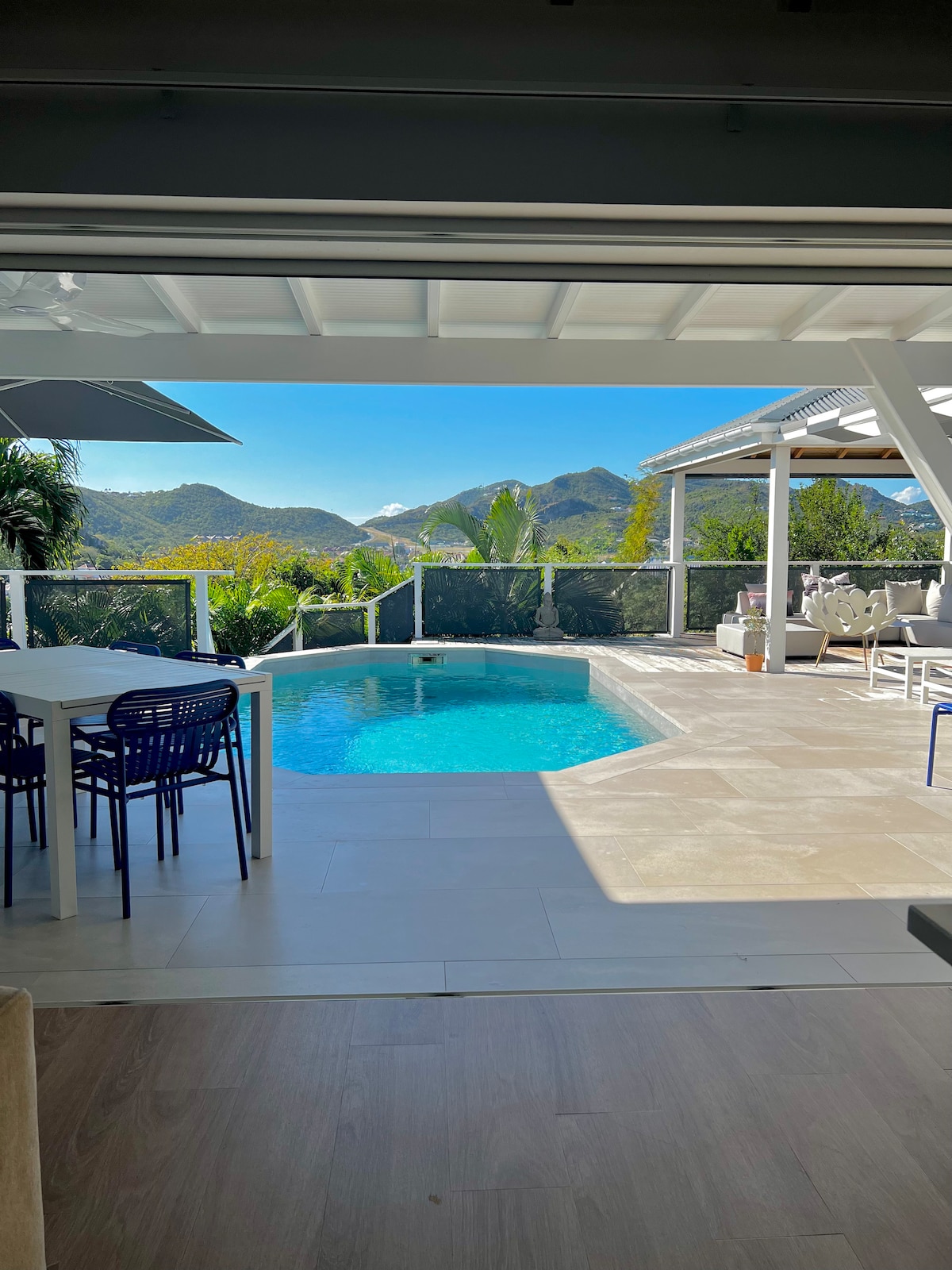 An inviting outdoor terrace features a swimming pool surrounded by sunlit tiles. Comfortable seating is positioned nearby, with a dining table hosted under a shaded area. Lush greenery and distant mountains create a scenic backdrop, enhancing the overall tranquility of the space.