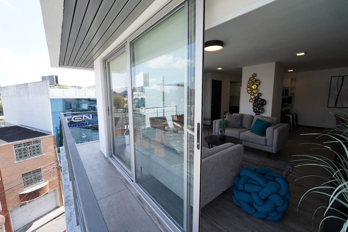 A spacious terrace features floor-to-ceiling glass doors that enhance the connection to the indoor living area. A modern seating arrangement is visible, complemented by decorative elements. The surrounding cityscape is partially visible beyond the railing.