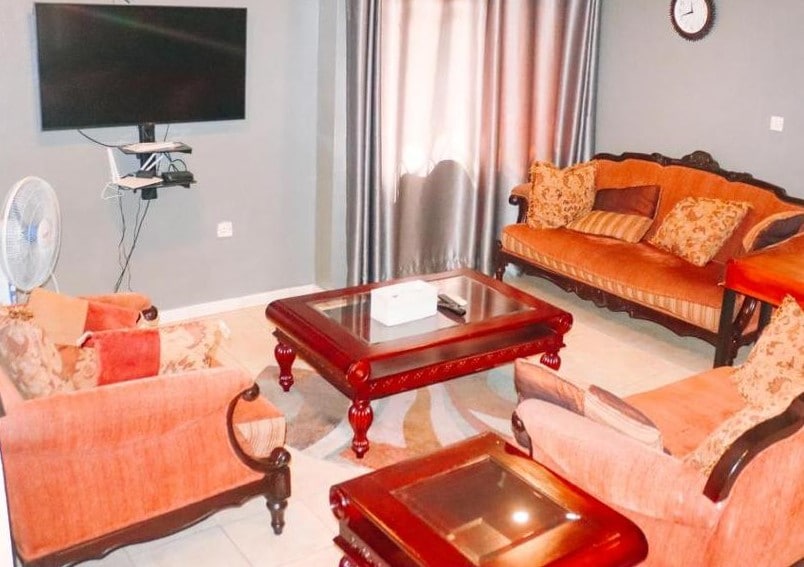 A comfortable living area is presented, featuring a flat-screen television mounted on the wall. Soft seating is provided by a couch and armchairs adorned with decorative cushions. A wooden coffee table occupies the center, surrounded by a light-colored area rug.