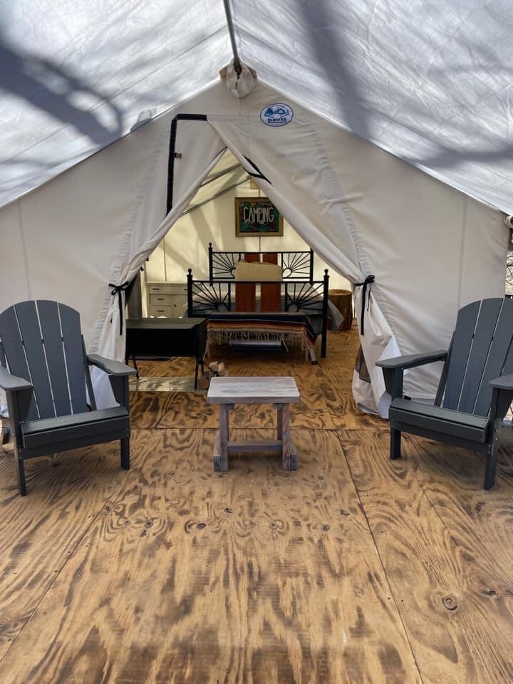 The entrance of a wall tent reveals a cozy interior with a queen bed, dresser, and wood-burning stove. Two black Adirondack chairs are placed outside on a wooden porch, accompanied by a small table. Natural light brightens the space, enhancing the serene environment.