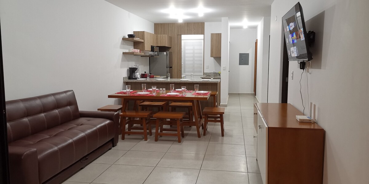 A modern living space features a dark brown sofa, a wooden dining table with seating for six, and a well-equipped kitchen. Natural light brightens the area, highlighting the simple cabinetry and appliances. A flat-screen TV is mounted on the wall, providing a functional entertainment option.