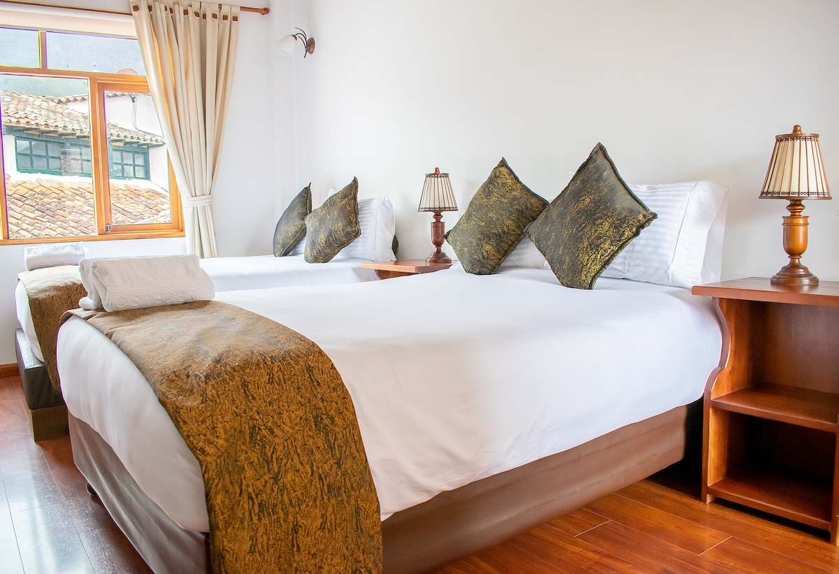 A bright and airy bedroom is presented, featuring two spacious beds adorned with decorative pillows. Wooden bedside tables with lamps are positioned on either side of the beds. Natural light streams in through a large window with sheer curtains, illuminating the warm wood flooring.