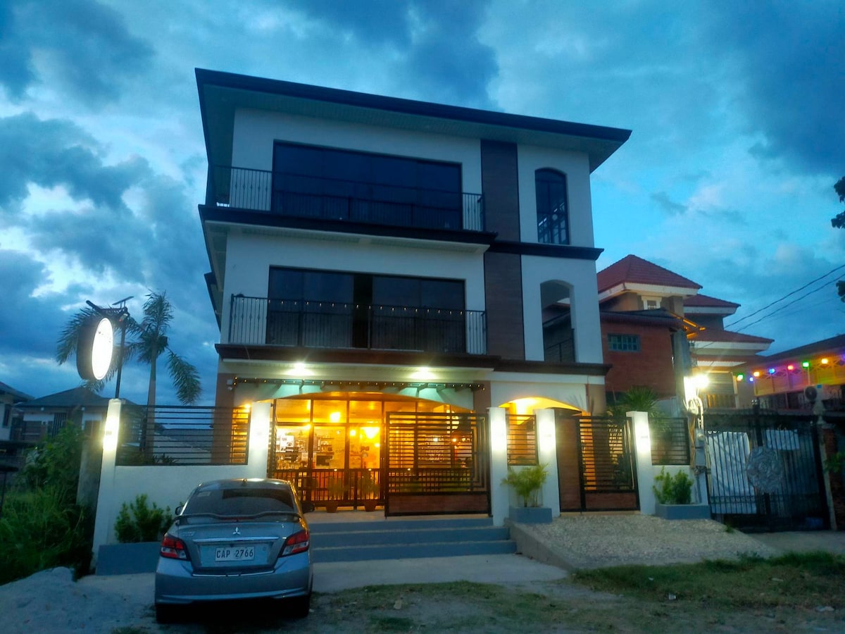 The exterior of a three-story building is depicted, featuring large windows on each level and a balcony on the upper floor. A car is parked in front, while the surrounding area is landscaped with a stone pathway and greenery. The sky is overcast, enhancing the evening ambiance.