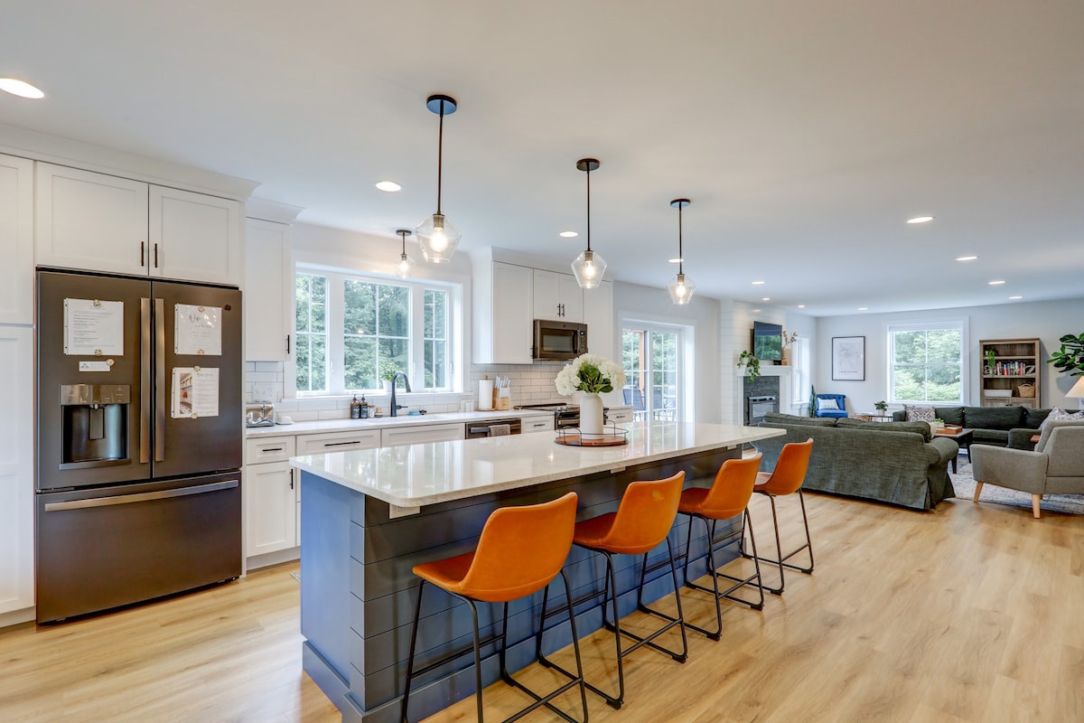 An open-concept kitchen featuring a large island with seating for four. The kitchen is equipped with stainless steel appliances and white cabinetry. Natural light enters through windows, illuminating the dining and living area in the background, which is furnished with comfortable seating and greenery.