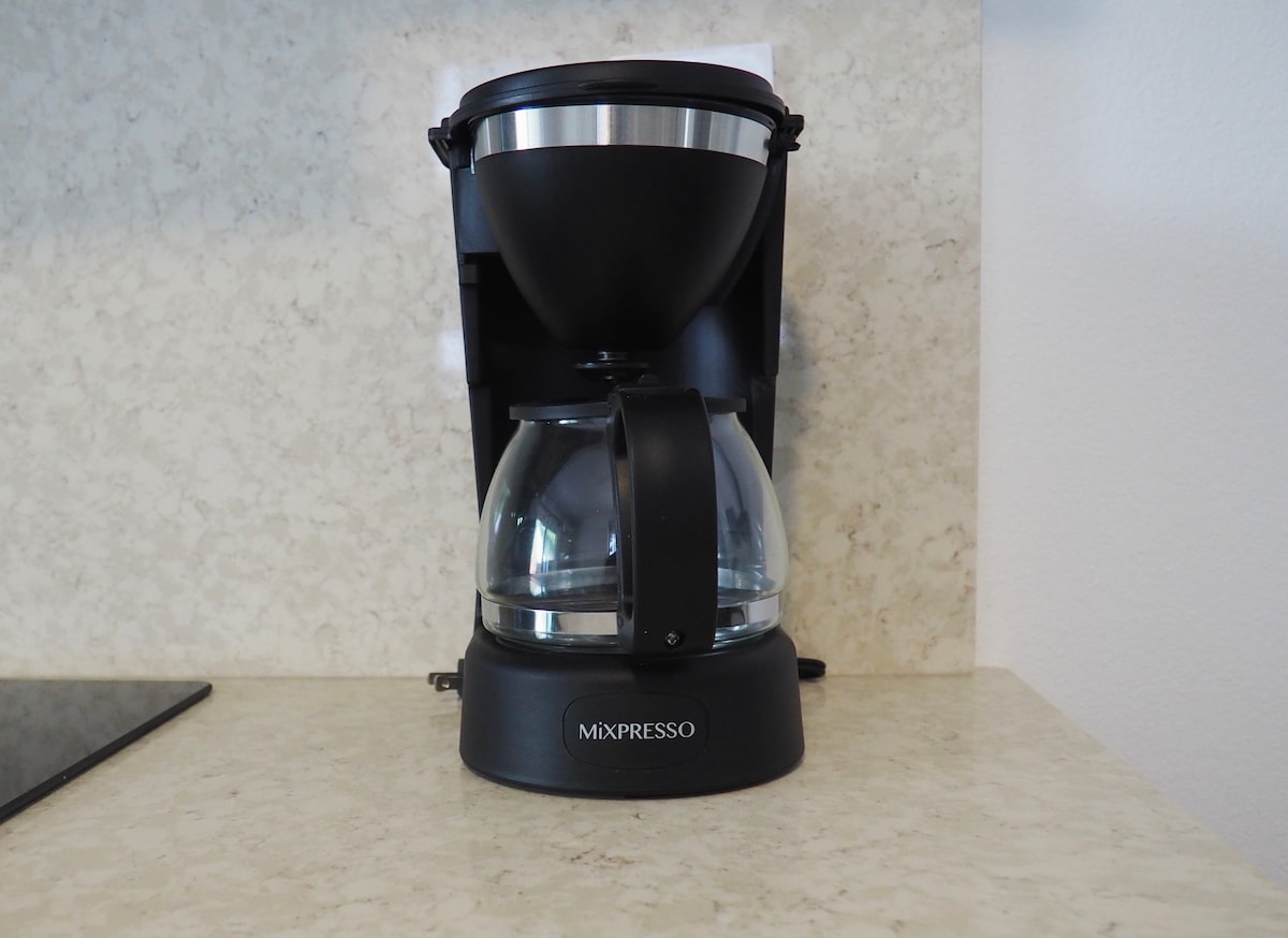 A coffee maker with a sleek black design is placed on a light-colored countertop. The glass carafe is visible, showcasing a rounded shape, and the machine features a stainless steel top. The background is a neutral-colored wall, enhancing the kitchen's modern vibe.