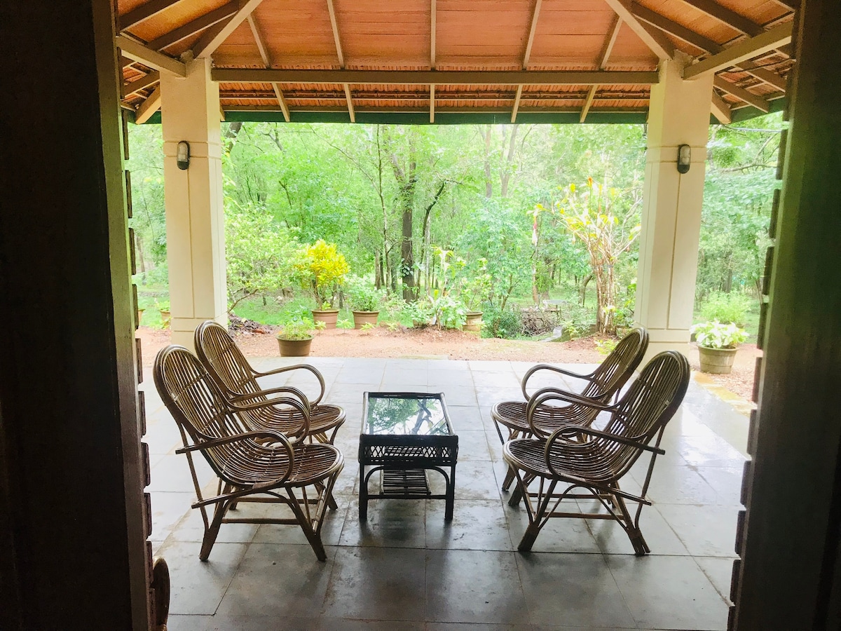 A spacious outdoor seating area is framed by wooden beams, featuring six rattan chairs arranged around a glass table. Lush greenery and blooming plants surround the space, creating a serene and inviting atmosphere. Natural light filters through the open structure, enhancing the connection to the outdoors.