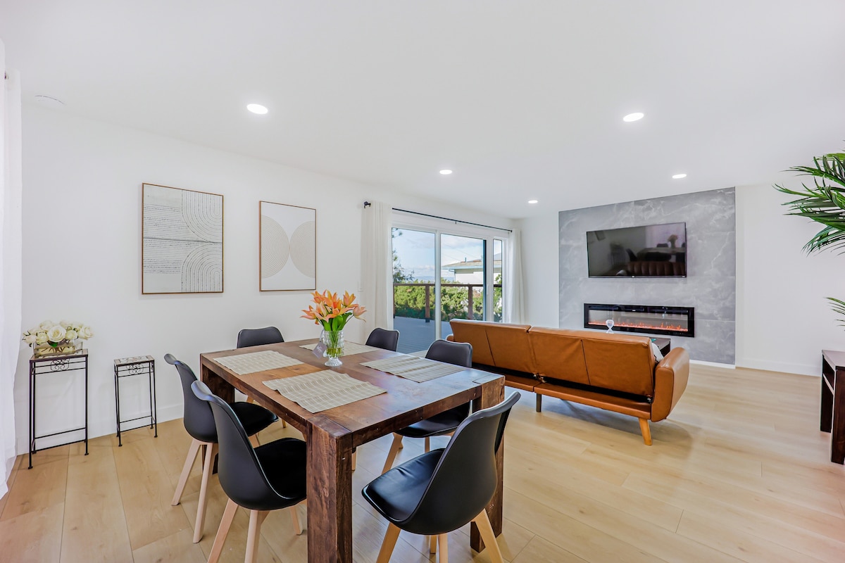 A modern dining area features a wooden table with seating for six guests, accompanied by stylish black chairs. Natural light brightens the space, highlighting a comfortable leather sofa and a wall-mounted television. A decorative plant adds a touch of greenery to the room.