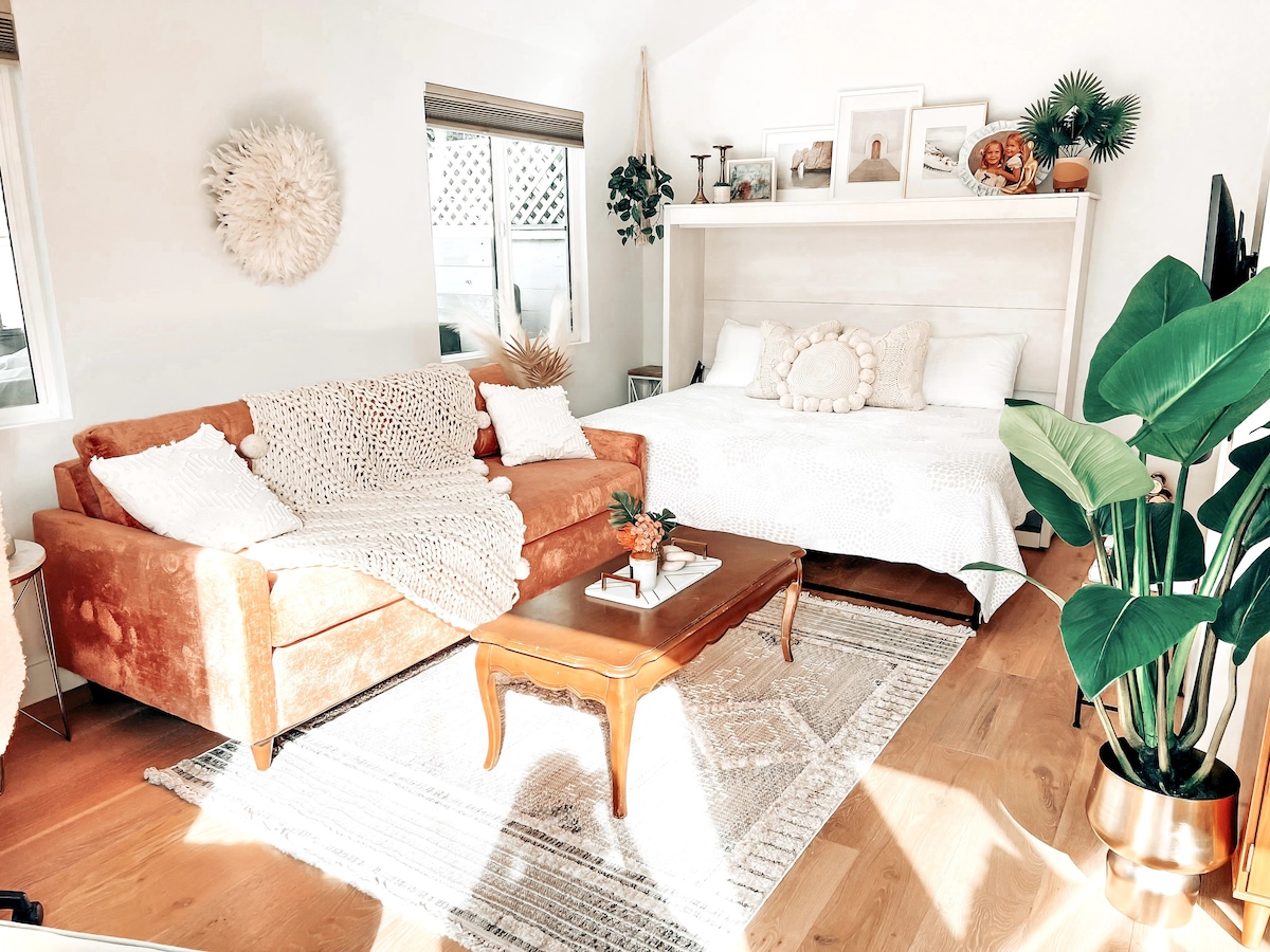 A modern living space features a spacious sofa with a textured throw and a decorative table. A double murphy bed with a light textile design is folded up against the wall. Natural light enters through two windows adorned with simple window treatments, complementing the warm wood flooring.