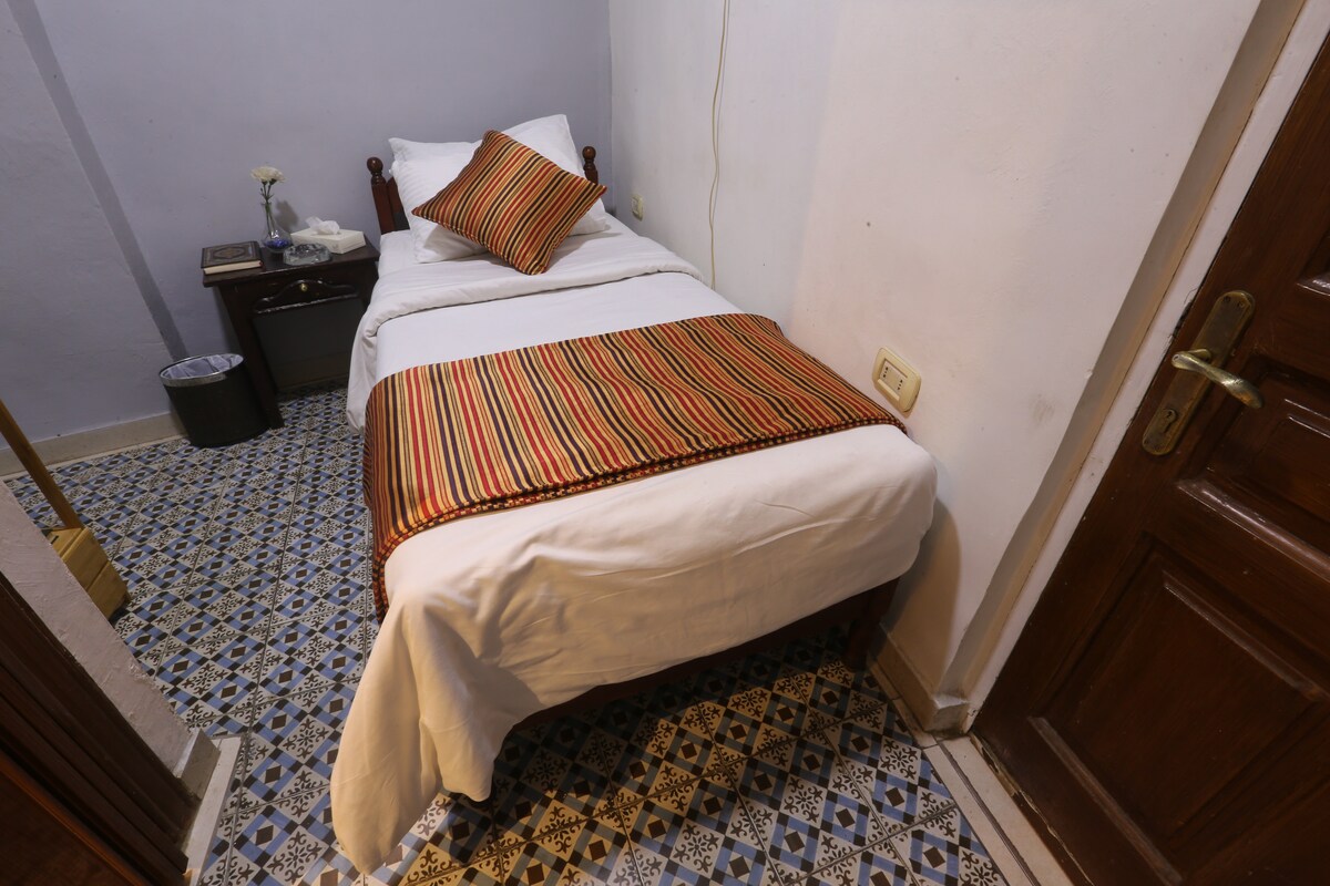 A compact bedroom features a twin bed with a colorful striped throw draped across the foot. A decorative pillow adds a touch of color. A nightstand is positioned beside the bed, with a small vase and a wastebasket nearby. The tiled floor exhibits a patterned design.
