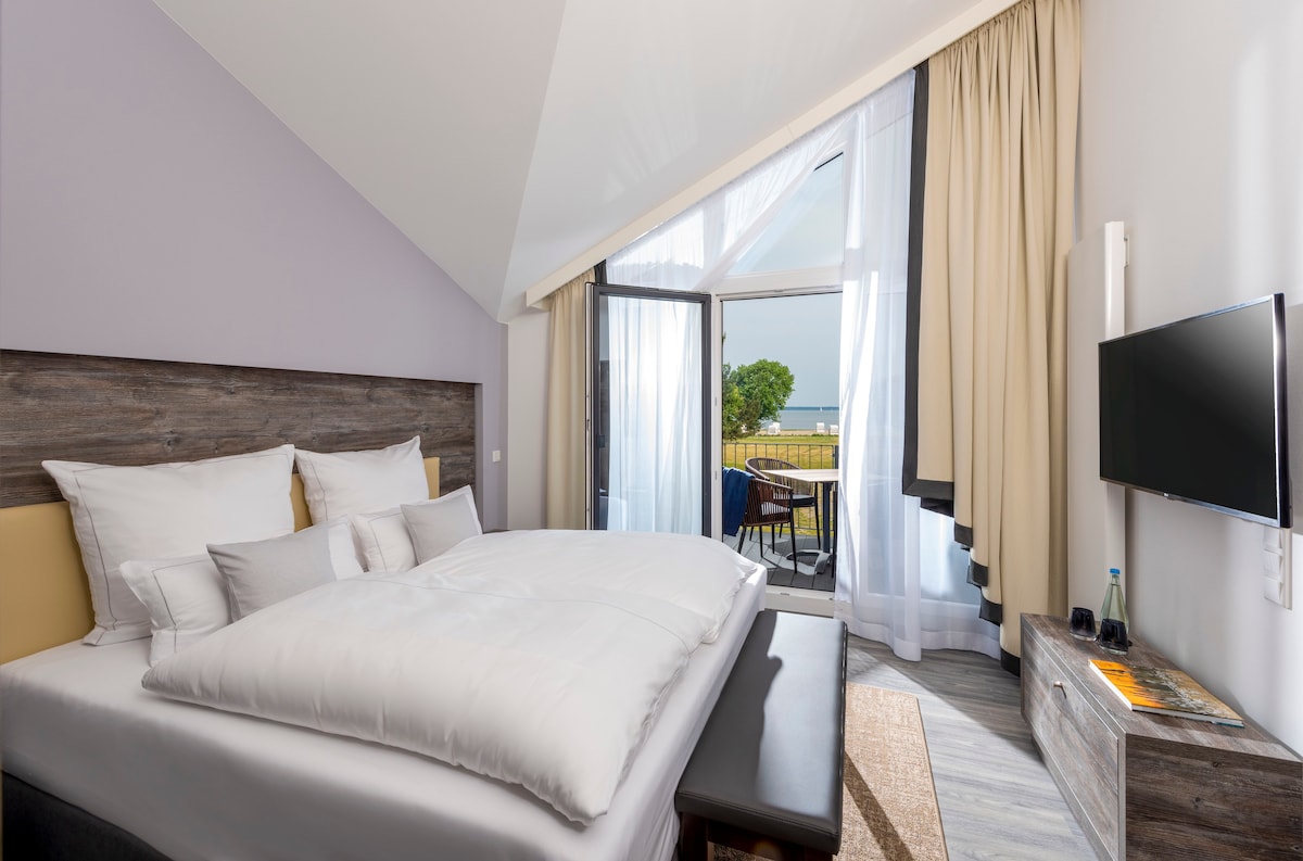 A spacious sleeping area features a double bed with plush bedding. Floor-to-ceiling windows allow natural light and provide access to a private balcony. Light-colored curtains frame the windows, while a flat-screen television and seating area complete the room's inviting layout.