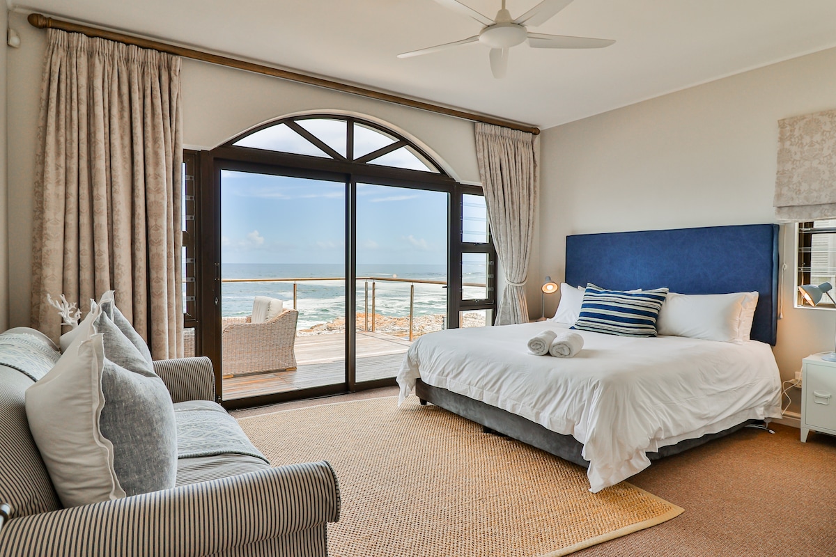 The bright bedroom features a spacious bed with a blue upholstered headboard. Large glass doors provide views of the ocean and access to a deck. A cozy seating area is present, complemented by textured curtains and a natural fiber rug underfoot.