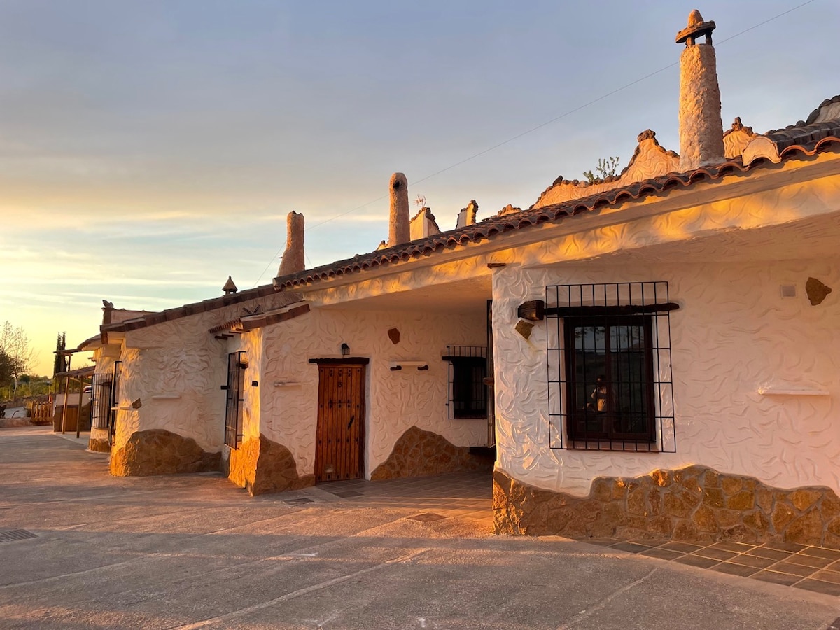 A unique cave dwelling with a rustic exterior is bathed in warm sunset light. The structure features a textured façade with natural stone elements, wooden doors, and traditional chimney details. Windows are framed with black bars, enhancing the characteristic look of the residence.
