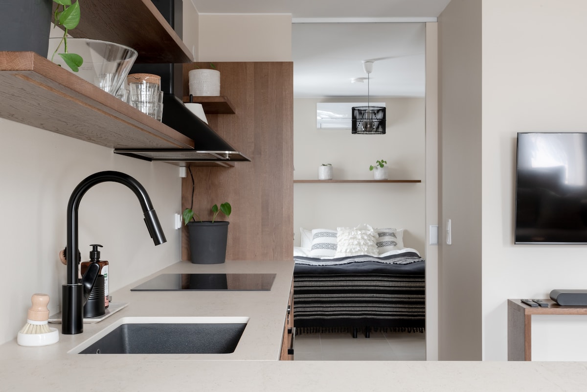 A modern kitchen area is displayed with sleek cabinetry and an integrated sink. A minimalist design is highlighted by a black faucet and subtly arranged kitchenware. In the background, a cozy sleeping space is visible, featuring a bed dressed in neutral tones and accented with soft textures.