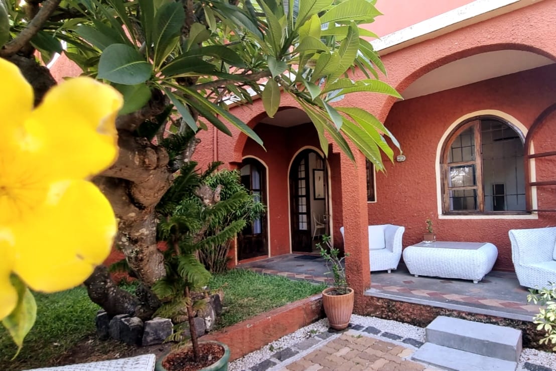 An inviting outdoor area features a flower-bearing tree and lush greenery. Two white seating arrangements are positioned on a stone patio, framed by arched doorways leading to the interior. The warm tones of the building’s exterior complement the tranquil garden setting.