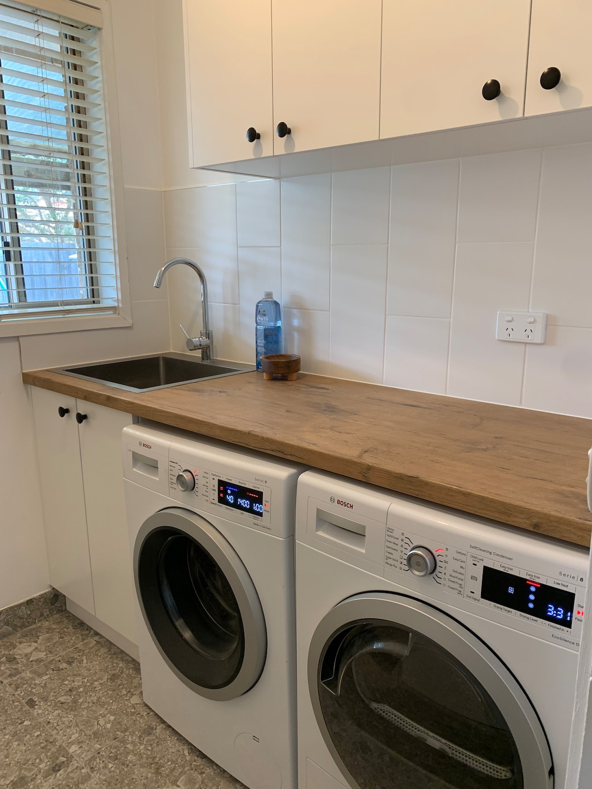A modern laundry area is equipped with a stacked washer and dryer, both featuring clear control panels. A wooden countertop provides additional workspace, and a stainless steel sink is positioned adjacent to the appliances. Natural light enters through a nearby window, illuminating the space.