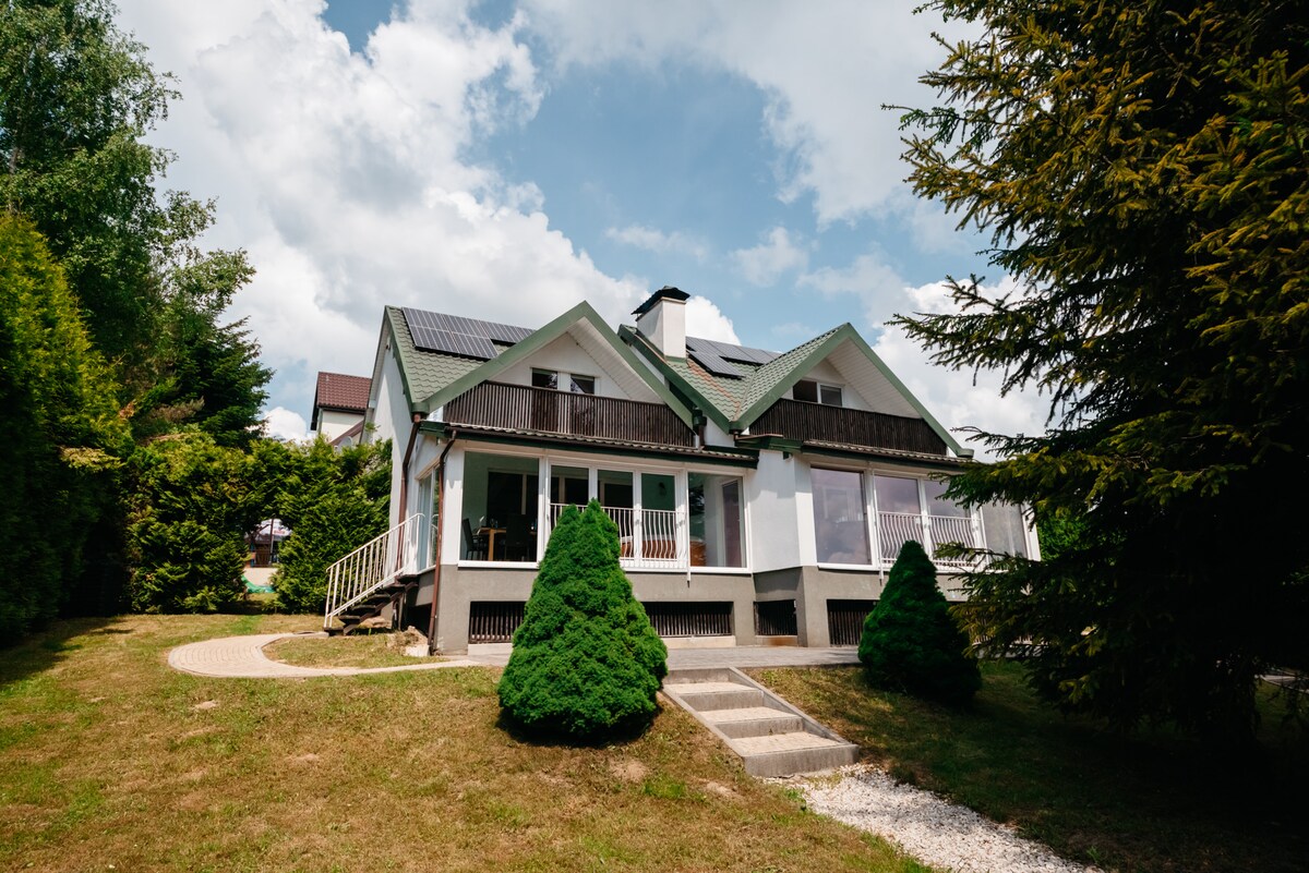 The exterior of a two-story house is depicted, featuring a sloping green roof and large windows. A walkway leads to a covered porch, surrounded by neatly trimmed shrubs. Lush greenery and a gravel path enhance the setting of the home, creating a welcoming outdoor space.