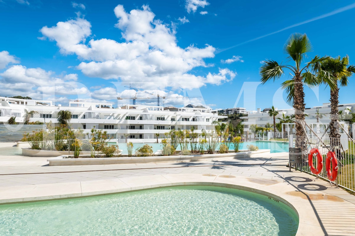 A community swimming pool area is visible, featuring a curved design and surrounded by decorative plants and palm trees. The clear blue sky and white clouds reflect a bright atmosphere. The nearby building exhibits a modern architectural style, enhancing the overall aesthetic of the setting.